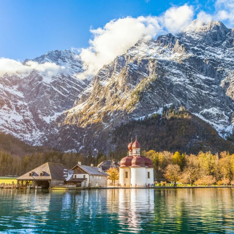 A person in traditional attire with a feathered headdress stands against a backdrop of large tree roots. The outfit includes a brown fringed garment and ornamental accessories. The setting appears to be a forest with dense wooden textures and hints of green foliage in the background.